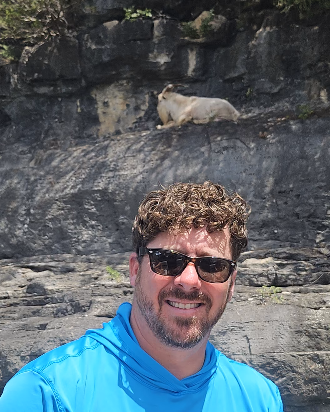 Captain Ryan Cash below a limestone bluff on Table Rock, with a goat resting on the ledge above him.